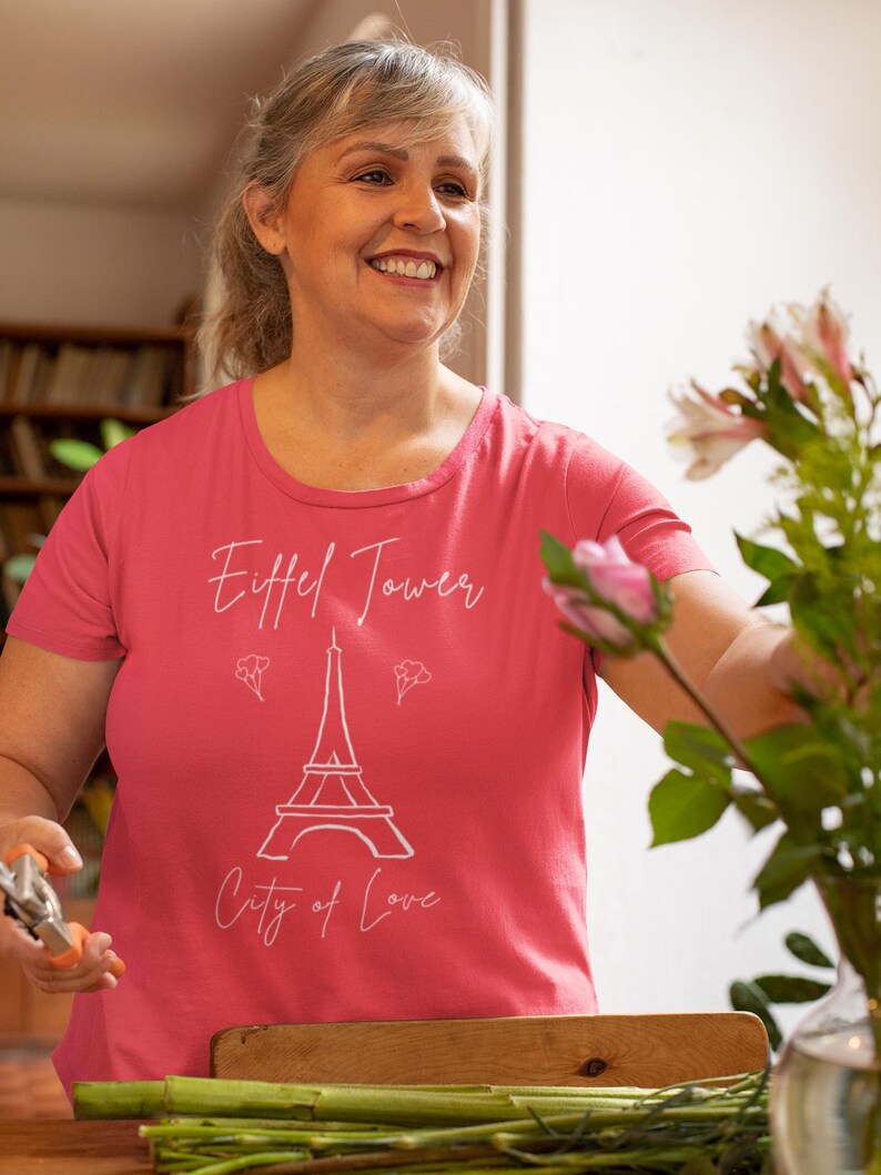 Op de afbeelding: Een koraalkleurig T-shirt met een witte afbeelding van de Eiffeltoren en de woorden "Eiffel Tower" en "City of Love". Het T-shirt heeft een ronde hals en korte mouwen. De afbeelding toont een lachende persoon. Het T-shirt is helder koraal van kleur.