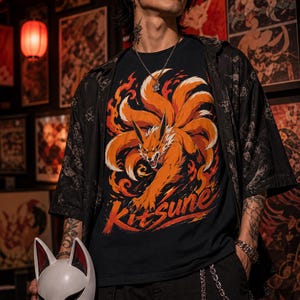 K&ouml;nnte beinhalten: Ein schwarzes T-Shirt mit einer Grafik eines orangefarbenen neunschw&auml;nzigen Fuchses, umgeben von Flammen, mit dem darunter gedruckten Text Kitsune. Das T-Shirt wird unter einem schwarzen kurz&auml;rmeligen Hemd getragen und eine wei&szlig;e Fuchsmaske.