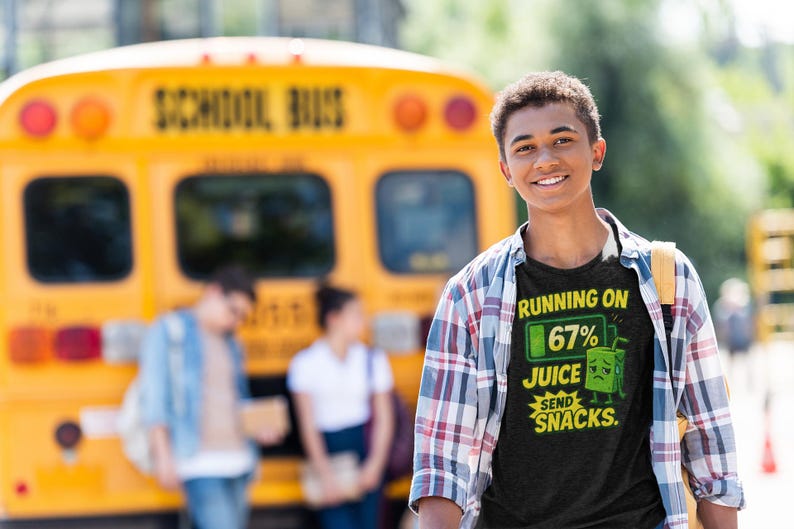 Pu&ograve; includere: Una persona indossa una maglietta nera con la scritta "Running on 67% Juice, Send Snacks". La maglietta presenta una grafica di un cartone di succo con una faccia triste. Sullo sfondo c'&egrave; un autobus scolastico giallo.