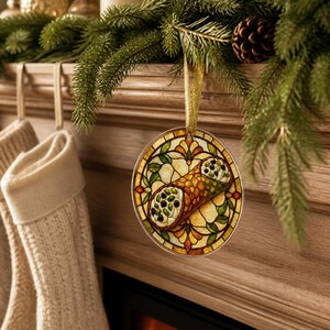 May include: A round, flat ornament featuring a stained-glass style illustration of a cannoli with a golden-brown pastry shell and white cream filling with dark chocolate chips, suspended by a thin gold ribbon against a festive holiday backdrop.