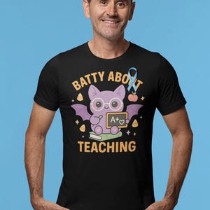 K&ouml;nnte beinhalten: Schwarzes T-Shirt mit einer Cartoon-Fledermaus mit Brille, die eine Tafel mit einem A+ h&auml;lt. Der Text "BATTY ABOUT TEACHING" ist in Gold gehalten, mit einem blauen Band und einer Apfelgrafik. Ein lustiges Design f&uuml;r Lehrer.