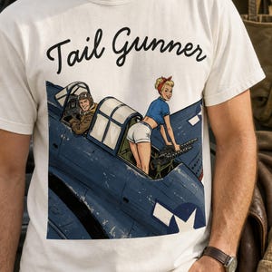May include: A cream-colored t-shirt featuring a graphic illustration of a blue military aircraft with a pilot in the cockpit and a pin-up style figure operating a machine gun. The text Tail Gunner is printed in black script above the illustration.