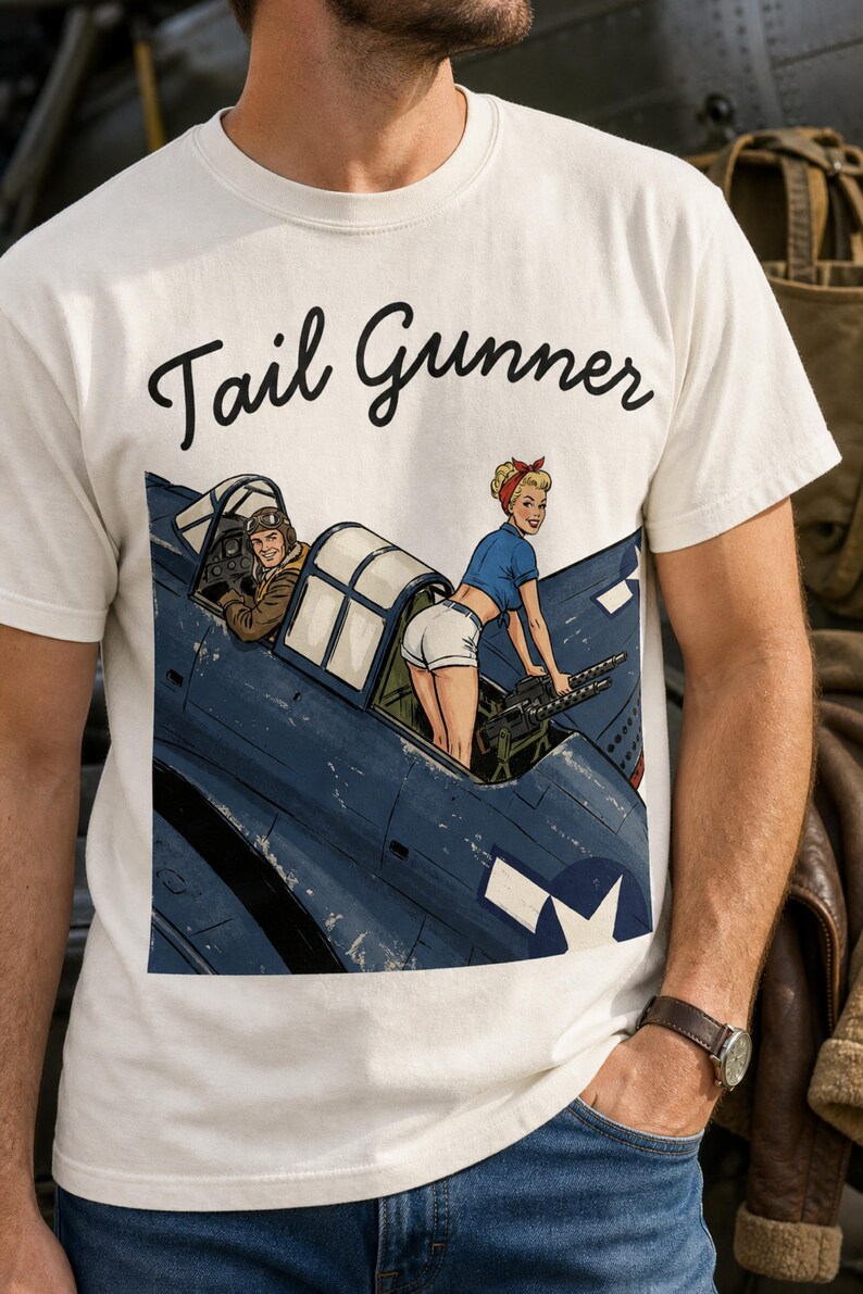 May include: A cream-colored t-shirt featuring a graphic illustration of a blue military aircraft with a pilot in the cockpit and a pin-up style figure operating a machine gun. The text Tail Gunner is printed in black script above the illustration.