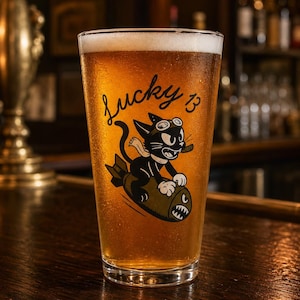 May include: A clear glass pint beer mug filled with golden amber liquid and white foam. The glass features a black graphic of a cartoon cat wearing goggles and smoking a cigar while riding a bomb, with the text Lucky 13 printed above the cat.