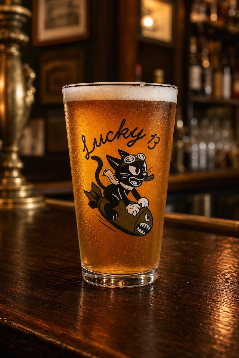 May include: A clear glass pint beer mug filled with golden amber liquid and white foam. The glass features a black graphic of a cartoon cat wearing goggles and smoking a cigar while riding a bomb, with the text Lucky 13 printed above the cat.