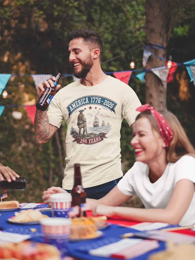 May include: A cream-coloured short-sleeve t-shirt featuring a graphic print with the text AMERICA 1776-2026 and 250 YEARS. The design includes a historical figure standing on a shore with sailing ships in the background, framed by banners.