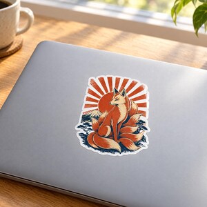 May include: A die-cut sticker featuring a multi-tailed orange fox sitting in front of a large red sun and a mountain silhouette. The design incorporates stylized blue waves and dark foliage at the base, rendered in a graphic illustration style.