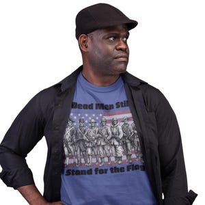 May include: A blue t-shirt featuring a graphic of seven skeleton soldiers in various historical military uniforms standing in a line. Behind the skeletons is a faded American flag. The text reads Dead Men Still Stand for the Flag.