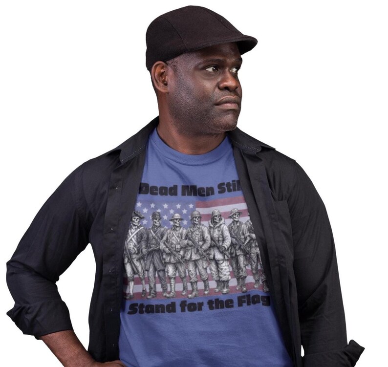 May include: A blue t-shirt featuring a graphic of seven skeleton soldiers in various historical military uniforms standing in a line. Behind the skeletons is a faded American flag. The text reads Dead Men Still Stand for the Flag.