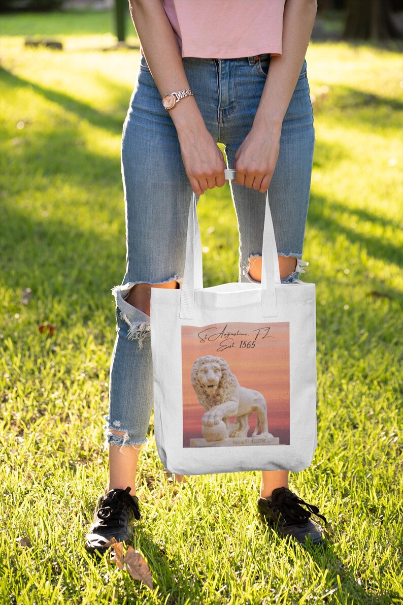 May include: White tote bag with a lion statue graphic and the text "St. Augustine, FL Est. 1565". The image has a sunset background. The bag is held by a person wearing blue jeans and black trainers.