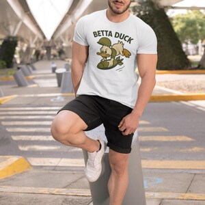 May include: White t-shirt featuring a cartoon duck graphic and the text "Betta Duck". The duck is wearing a military helmet and riding a bomb. The person is wearing black shorts and white trainers.