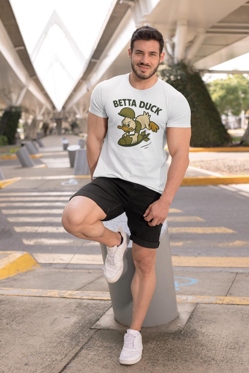 May include: White t-shirt featuring a cartoon duck graphic and the text "Betta Duck". The duck is wearing a military helmet and riding a bomb. The person is wearing black shorts and white trainers.