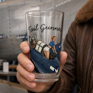 May include: A clear glass pint tumbler featuring a graphic illustration of a blue military aircraft with a person in a blue shirt and white shorts operating a machine gun, with the text Tail Gunner printed in black script above the illustration.