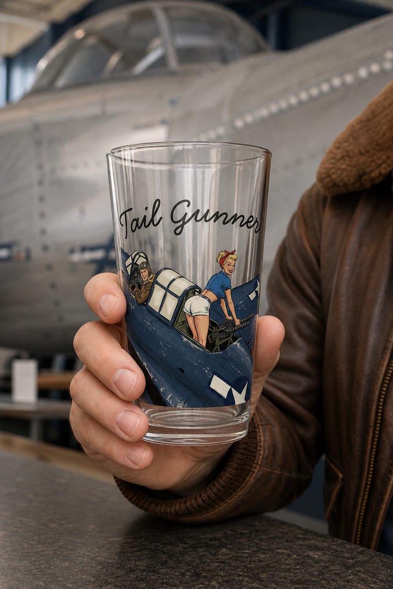 May include: A clear glass pint tumbler featuring a graphic illustration of a blue military aircraft with a person in a blue shirt and white shorts operating a machine gun, with the text Tail Gunner printed in black script above the illustration.