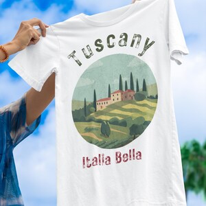 May include: White t-shirt featuring a circular graphic of a Tuscan landscape with rolling hills, cypress trees, and a villa. The text "Tuscany" and "Italia Bella" are printed on the shirt.