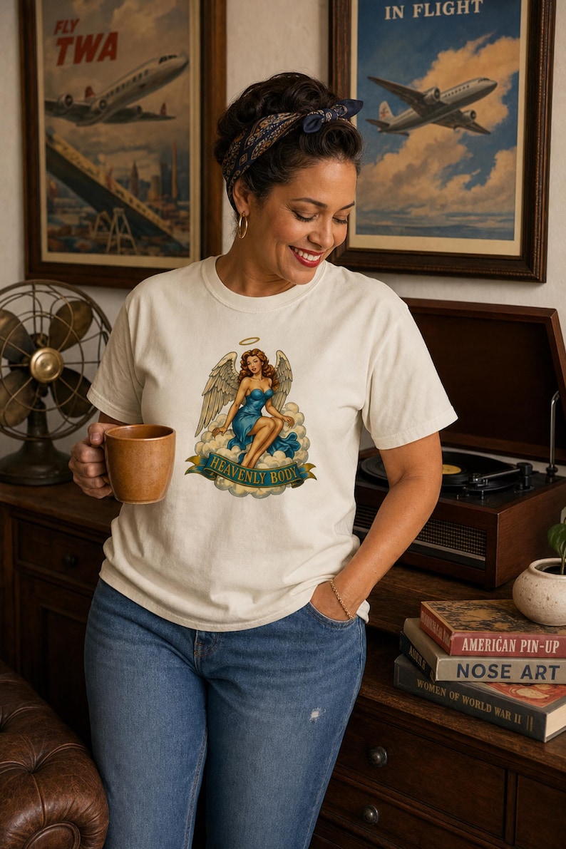 May include: An off-white short-sleeve t-shirt featuring a graphic of an angel with brown hair and a blue dress sitting on clouds. Below the angel, a blue banner contains the text "HEAVENLY BODY" in gold lettering. The shirt has a crew neckline.