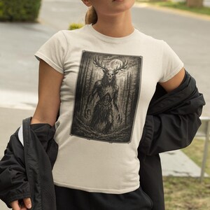 May include: A cream-colored short-sleeve t-shirt featuring a rectangular graphic print of a dark, skeletal creature with large antlers standing in a dense forest under a full moon. The illustration is rendered in shades of black, gray, and white.