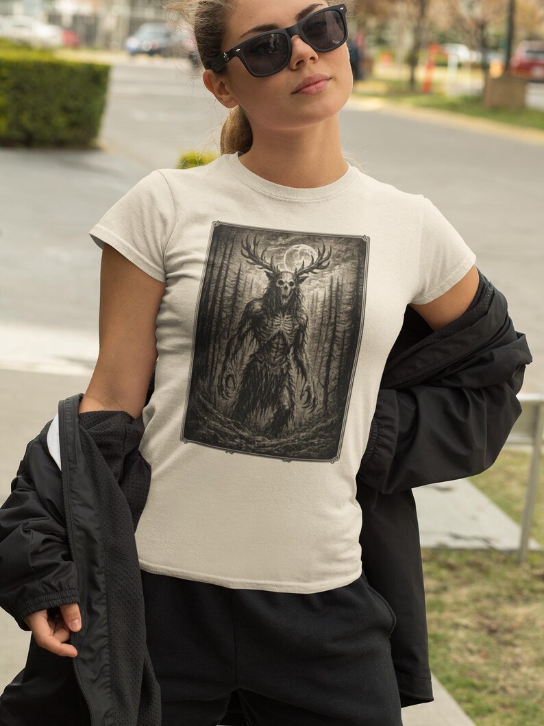 May include: A cream-colored short-sleeve t-shirt featuring a rectangular graphic print of a dark, skeletal creature with large antlers standing in a dense forest under a full moon. The illustration is rendered in shades of black, gray, and white.