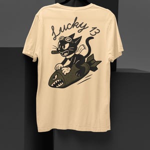 May include: Cream-colored t-shirt featuring a cartoon cat riding a bomb. The cat wears goggles and a cigar. The text "Lucky 13" is above the cat. The shirt is displayed on a black hanger.