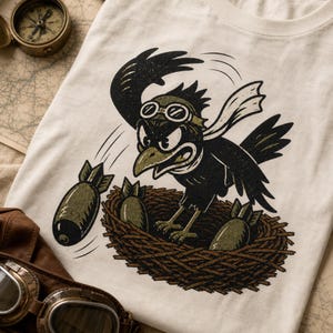 Puede incluir: Una camiseta color hueso con el gr&aacute;fico de un p&aacute;jaro negro con gafas de aviador en un nido. El p&aacute;jaro est&aacute; lanzando bombas de color verde oliva. La ilustraci&oacute;n utiliza un estilo de tinta desgastado con contornos negros y tonos verdes.