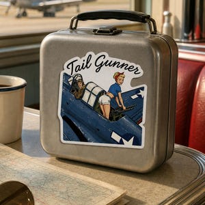 May include: A silver metal lunch box featuring a sticker with a blue military aircraft and a pin-up style character operating a machine gun. The sticker includes the text Tail Gunner in a black script font. The lunch box has a black handle.