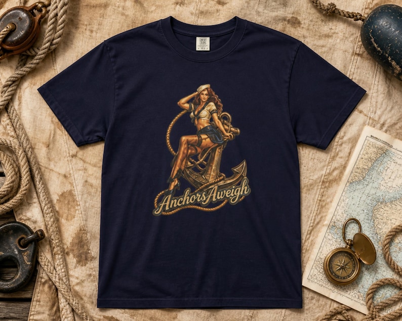 May include: A navy blue short-sleeve t-shirt featuring a graphic illustration of a person in a sailor outfit sitting on a large metal anchor. The text "Anchors Aweigh" is printed in a cursive font below the anchor, surrounded by a rope design.