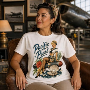 May include: A white short-sleeve t-shirt featuring a graphic print of a pin-up style sailor sitting on a torpedo next to a large open oyster shell with a pearl. The text Pacific Pearl is printed in blue script above the illustration.