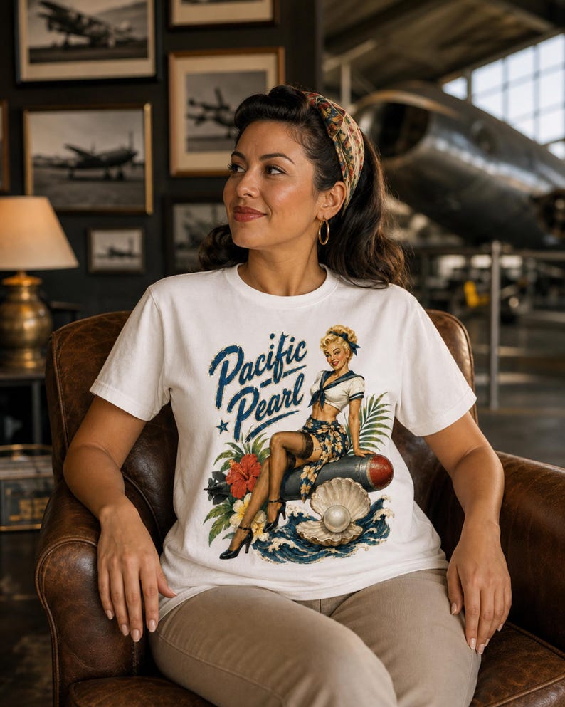 May include: A white short-sleeve t-shirt featuring a graphic print of a pin-up style sailor sitting on a torpedo next to a large open oyster shell with a pearl. The text Pacific Pearl is printed in blue script above the illustration.