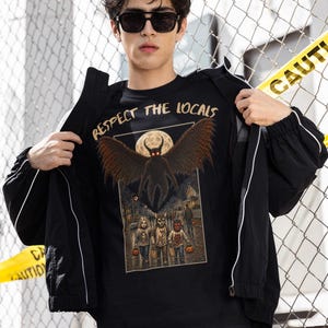 May include: A black t-shirt featuring a graphic print with the text "RESPECT THE LOCALS" above a winged creature with glowing red eyes and a full moon. Below, three figures in costumes stand on a street at night, each holding a pumpkin bucket.