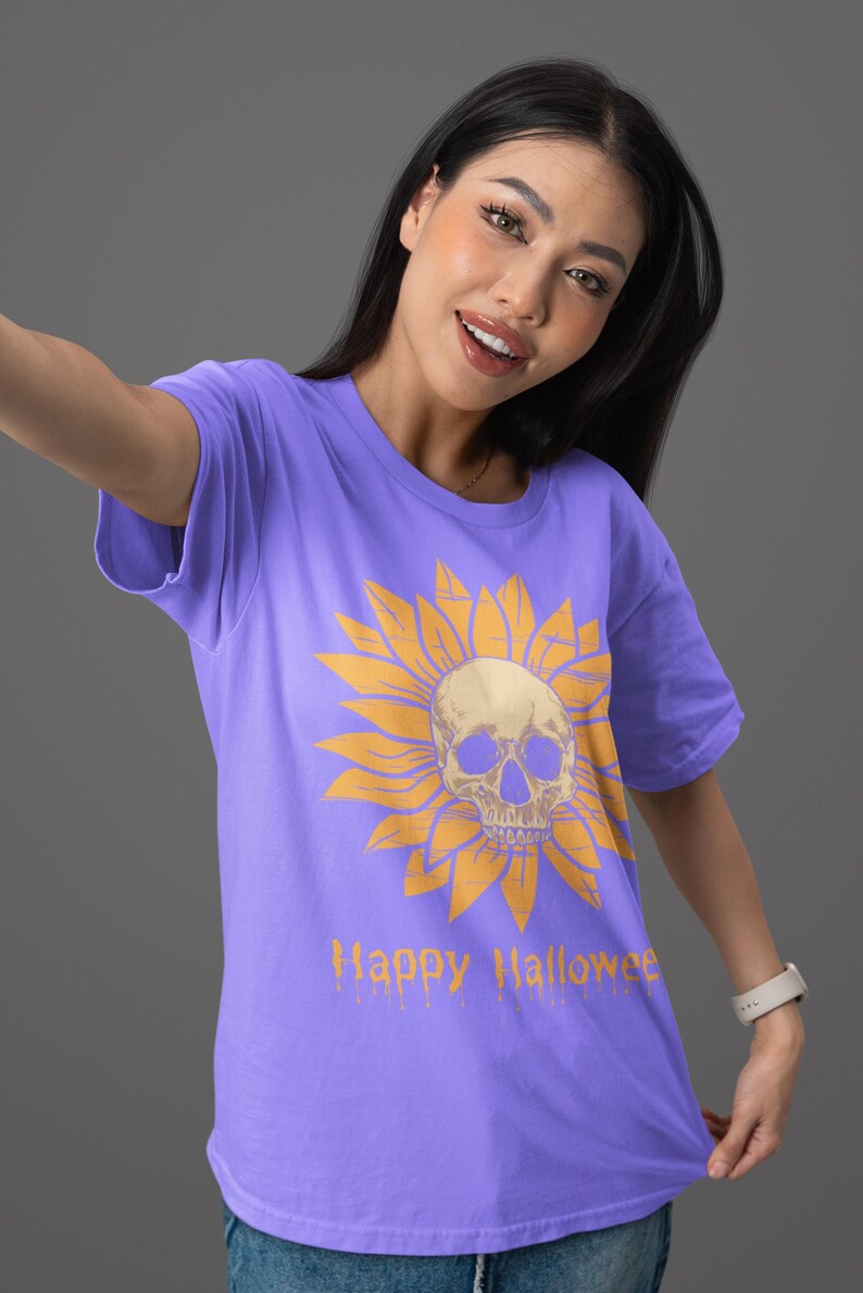 May include: A purple t-shirt featuring a yellow sunflower design with a skull in the center. The words "Happy Halloween" are printed in orange below the graphic. The shirt is a short-sleeved crew neck.