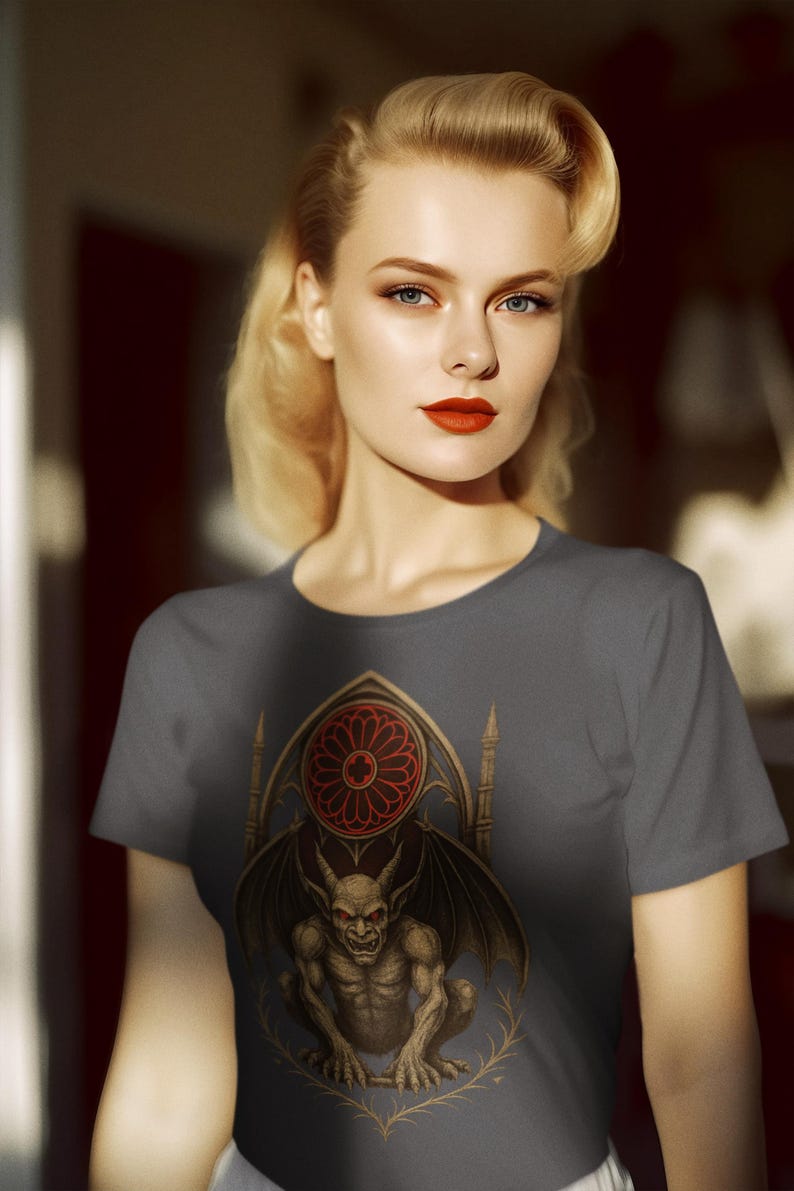May include: A gray t-shirt featuring a detailed illustration of a gargoyle with bat wings, set against a gothic window design with a red rose window. The shirt is worn by a person with blonde hair and red lipstick.