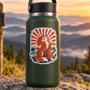 May include: A matte dark green insulated water bottle with a black lid and a sticker featuring a multi-tailed fox sitting in front of a red sunburst and a mountain silhouette. The sticker design uses orange, red, white, and blue color tones.