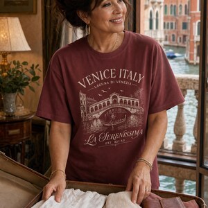 May include: A maroon short-sleeved t-shirt featuring a white graphic print of the Rialto Bridge in Venice, Italy. The text reads VENICE ITALY, LAGUNA DI VENEZIA, LA SERENISSIMA, and EST. 421 AD. The shirt has a crew neckline and a relaxed fit.