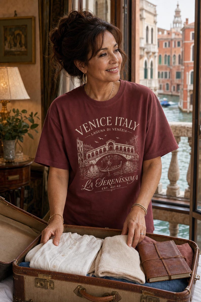 May include: A maroon short-sleeved t-shirt featuring a white graphic print of the Rialto Bridge in Venice, Italy. The text reads VENICE ITALY, LAGUNA DI VENEZIA, LA SERENISSIMA, and EST. 421 AD. The shirt has a crew neckline and a relaxed fit.