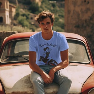 May include: Light blue t-shirt with the word "Bombshell" and a graphic of a woman riding a shark. The shirt is worn by a person sitting on the hood of a vintage red car.