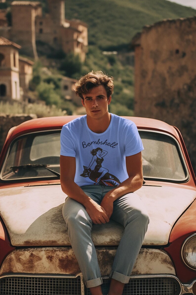 May include: Light blue t-shirt with the word "Bombshell" and a graphic of a woman riding a shark. The shirt is worn by a person sitting on the hood of a vintage red car.