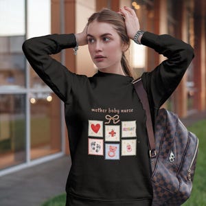 K&ouml;nnte beinhalten: Schwarzes Sweatshirt mit dem Text "mother baby nurse" und mehreren Briefmarken-Grafiken. Die Grafiken zeigen ein Herz, ein Kreuz, das Wort "nurse", Babyf&uuml;&szlig;e, einen Teddyb&auml;r und eine Flasche.