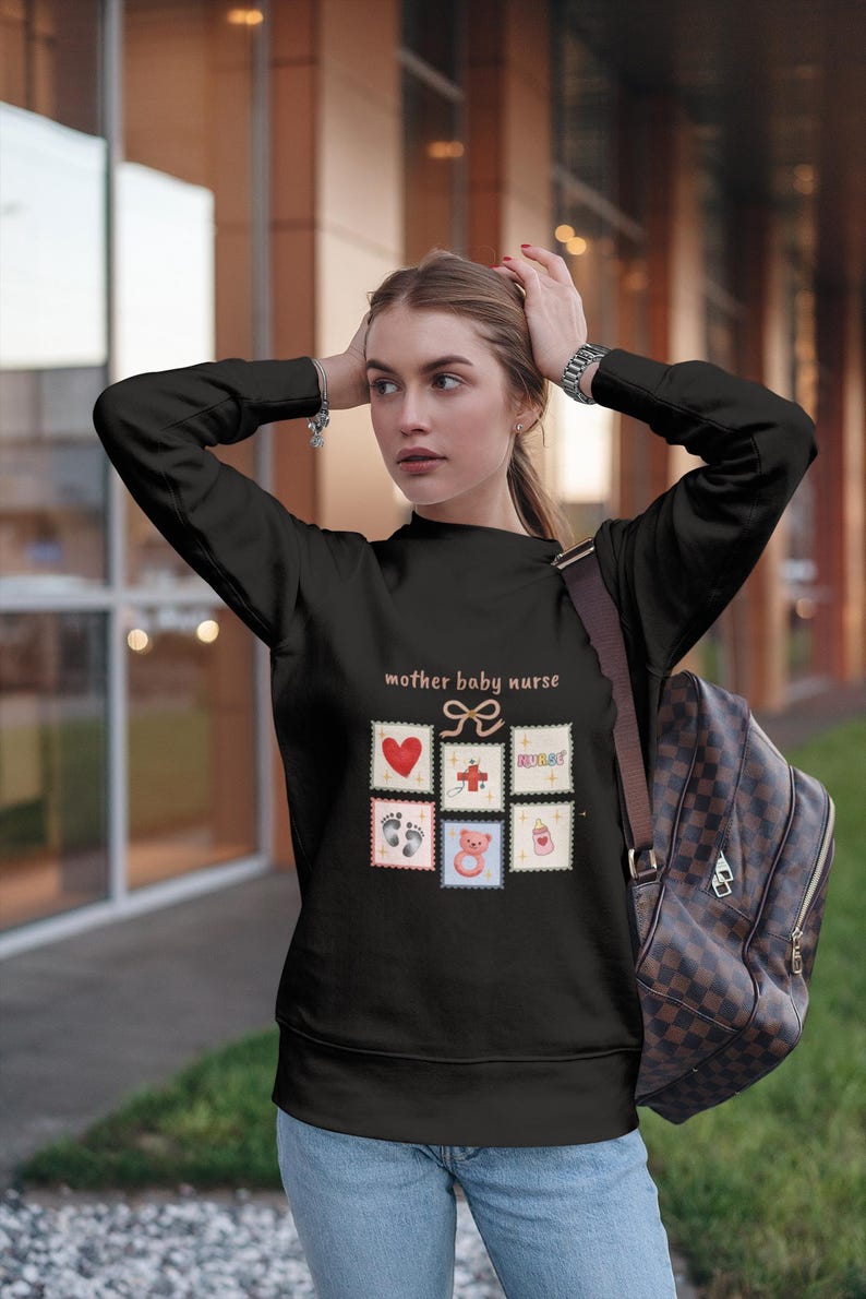 K&ouml;nnte beinhalten: Schwarzes Sweatshirt mit dem Text "mother baby nurse" und mehreren Briefmarken-Grafiken. Die Grafiken zeigen ein Herz, ein Kreuz, das Wort "nurse", Babyf&uuml;&szlig;e, einen Teddyb&auml;r und eine Flasche.