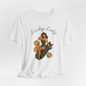 May include: A white short-sleeve t-shirt featuring a graphic of a woman in a green swimsuit sitting on a dark bomb, surrounded by three yellow dice. The text Lucky Lady is printed in a cursive grey font above the illustration on the front.
