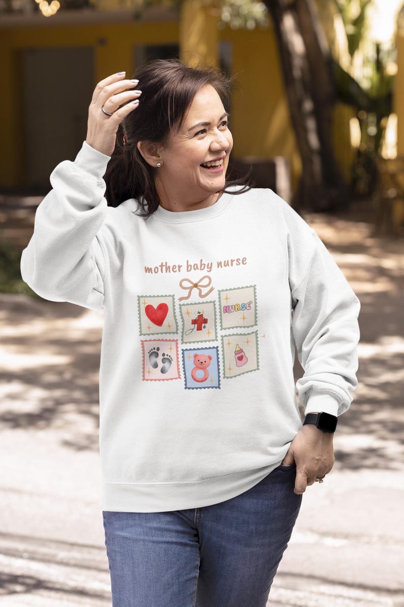 K&ouml;nnte beinhalten: Wei&szlig;es Sweatshirt mit dem Text "mother baby nurse" und mehreren Briefmarken-Grafiken. Die Grafiken zeigen ein rotes Herz, ein Kreuz, das Wort "nurse", Babyf&uuml;&szlig;e, einen Teddyb&auml;r und eine Babyflasche.