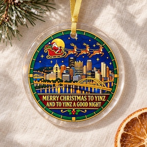 May include: A round clear acrylic ornament featuring a stained-glass style illustration of a city skyline at night with a bridge, Santa in a sleigh with reindeer, and the text MERRY CHRISTMAS TO YINZ AND TO YINZ A GOOD NIGHT, hung by a gold ribbon.