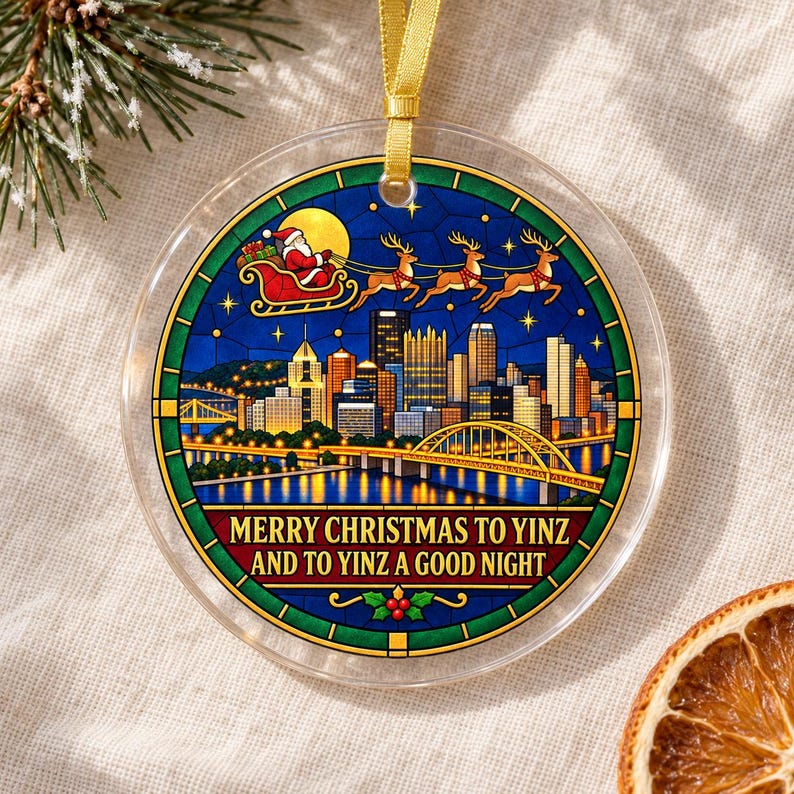 May include: A round clear acrylic ornament featuring a stained-glass style illustration of a city skyline at night with a bridge, Santa in a sleigh with reindeer, and the text MERRY CHRISTMAS TO YINZ AND TO YINZ A GOOD NIGHT, hung by a gold ribbon.