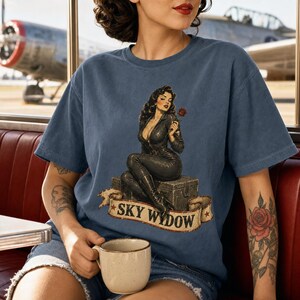 May include: A blue short-sleeve t-shirt featuring a graphic of a woman in a black jumpsuit sitting on a crate, holding a red rose. Below the figure, a banner displays the text SKY WIDOW in black capital letters, flanked by two red stars.