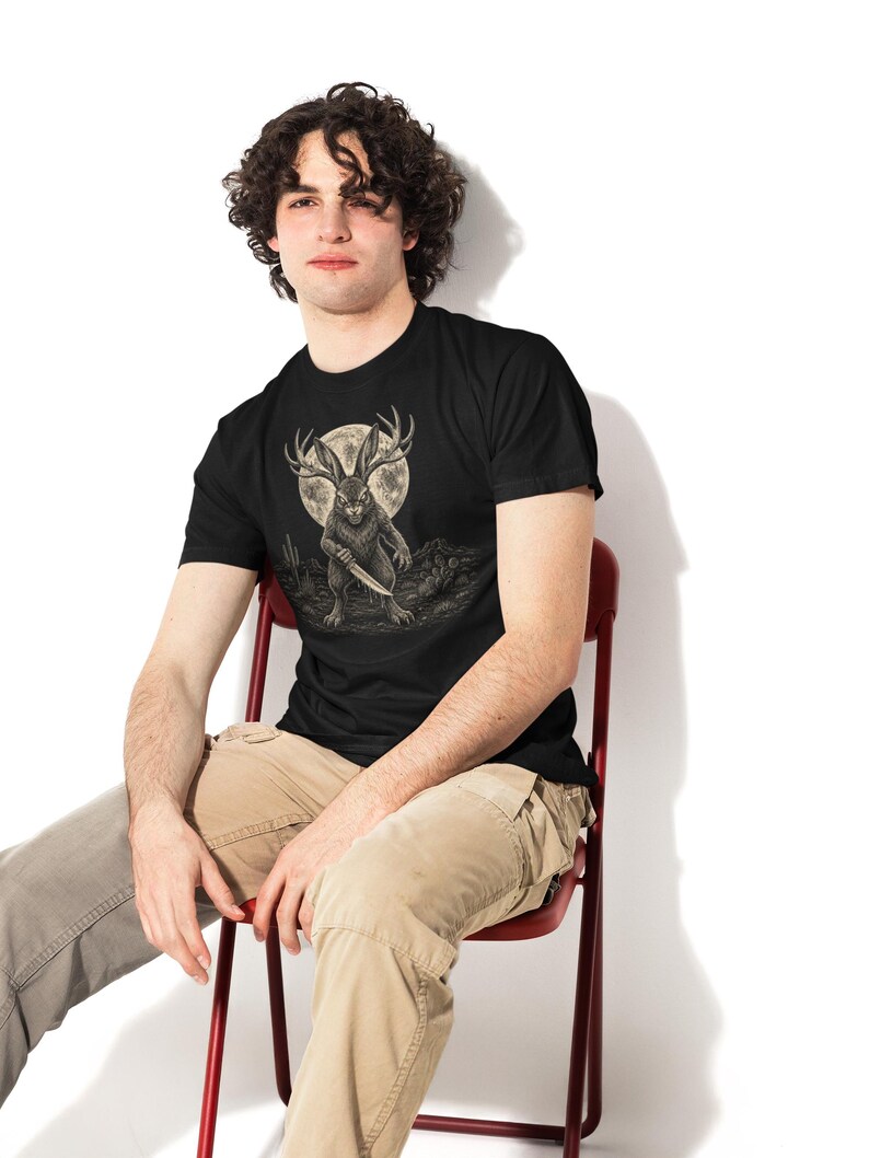 K&ouml;nnte beinhalten: Ein schwarzes T-Shirt mit kurzen &Auml;rmeln und einer grafischen Illustration eines Jackalopen, der ein Messer h&auml;lt. Das Wesen steht in einer W&uuml;stenlandschaft unter einem gro&szlig;en Vollmond. Das Design ist in Grau-, Wei&szlig;- und Schwarzt&ouml;nen gehalten.