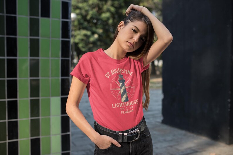 May include: A red t-shirt featuring the text "St. Augustine Lighthouse" with a lighthouse graphic. The shirt includes the text "Est. 1874 Florida". The model is wearing black jeans and a black belt.