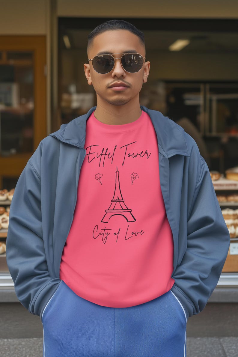 May include: A person wearing a pink t-shirt with a black Eiffel Tower graphic and the words 'Eiffel Tower City of Love'. The person is also wearing a blue jacket and sunglasses. The background is a storefront.