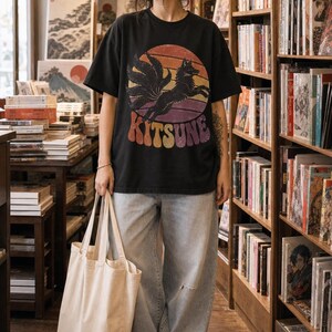 May include: A black short-sleeved t-shirt featuring a graphic of a multi-tailed fox leaping in front of a sunset circle with the text KITSUNE in a retro font, paired with light-wash blue denim jeans and a plain beige canvas tote bag.