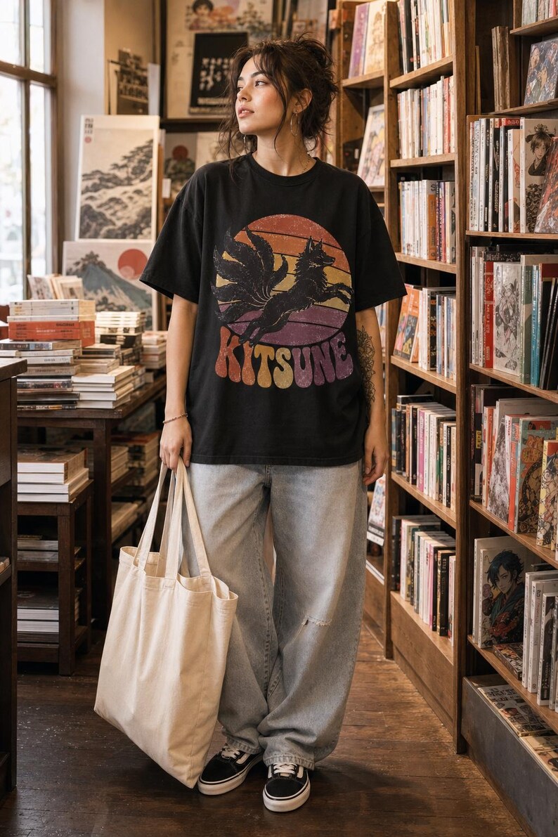 May include: A black short-sleeved t-shirt featuring a graphic of a multi-tailed fox leaping in front of a sunset circle with the text KITSUNE in a retro font, paired with light-wash blue denim jeans and a plain beige canvas tote bag.