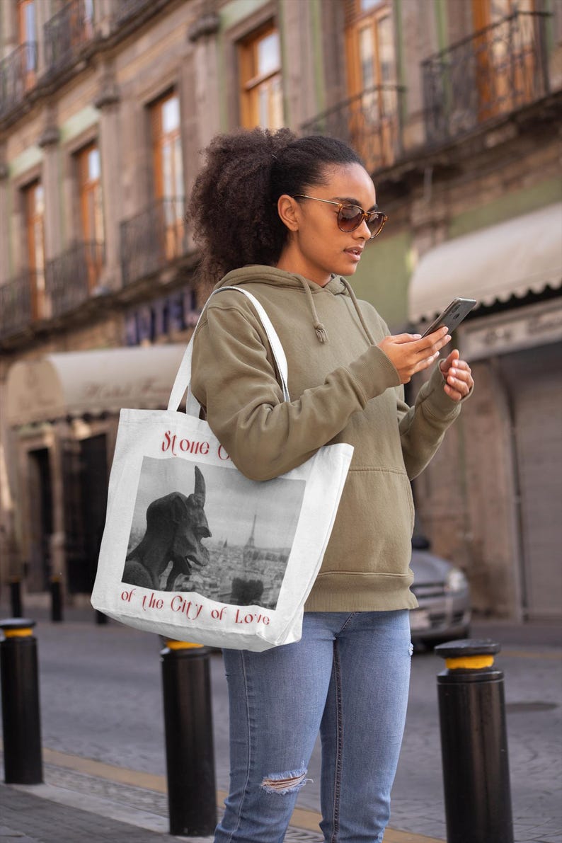 May include: A white tote bag with a black and white image of a gargoyle and the Eiffel Tower, with the text "Stone of the City of Love". The bag is carried by a person wearing a green hoodie and blue jeans.