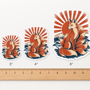 May include: Four die-cut stickers featuring a nine-tailed fox sitting before a red sun and mountain. Sizes are labeled 2, 3, 4, and 6 inches above a wooden ruler. The design includes orange, red, and dark blue tones with a white border.
